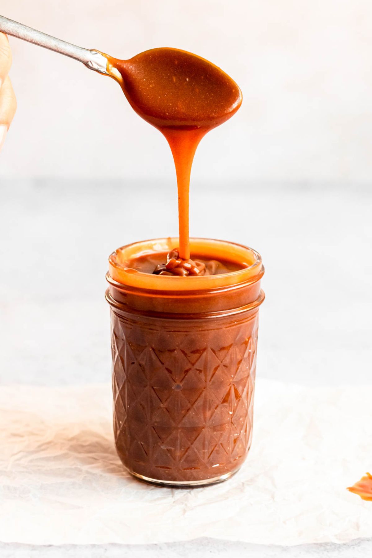 A spoon drizzling homemade salted caramel sauce into a glass mason jar against a light background.