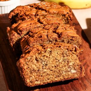 Sliced Biscoff banana bread drizzled with Biscoff spread on a wooden board alongside fresh bananas and a bowl of Biscoff cookies.