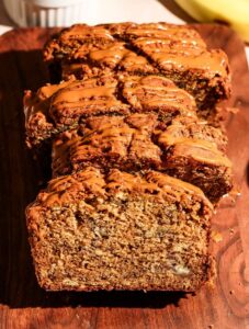 Close-up of sliced Biscoff banana bread drizzled with Biscoff spread, with a bowl of cookies and fresh bananas in the background.