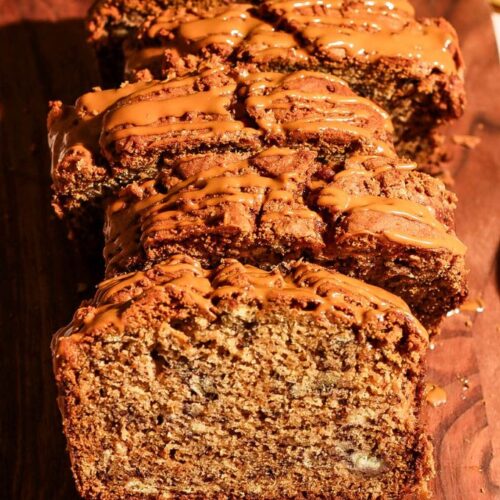 Close-up of sliced Biscoff banana bread drizzled with Biscoff spread, with a bowl of cookies and fresh bananas in the background.