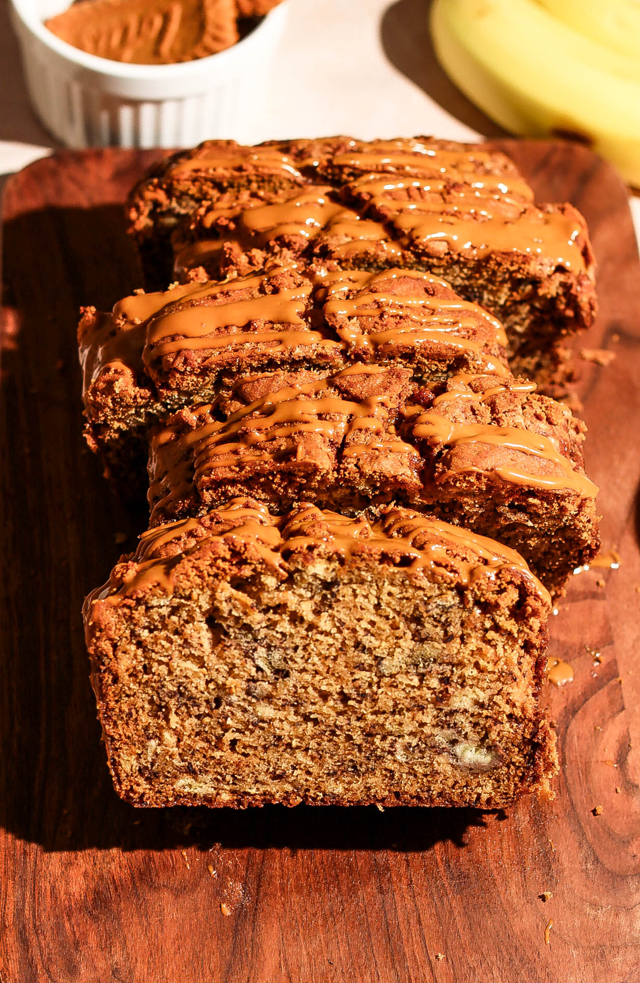 Close-up of sliced Biscoff banana bread drizzled with Biscoff spread, with a bowl of cookies and fresh bananas in the background.