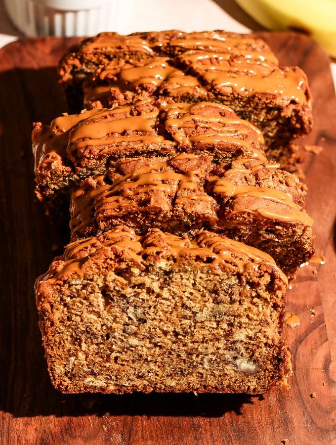 Close-up of sliced Biscoff banana bread drizzled with Biscoff spread, with a bowl of cookies and fresh bananas in the background.