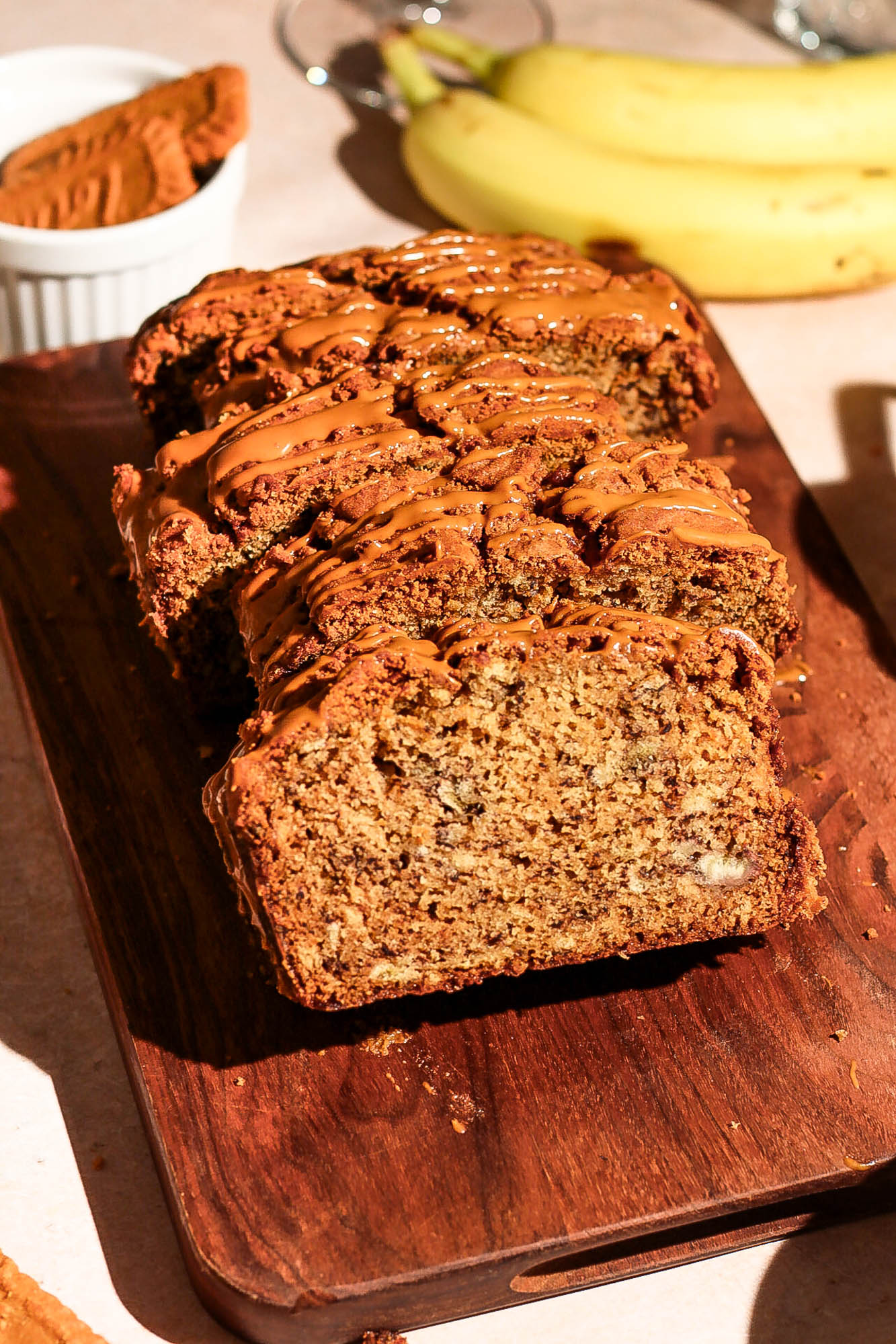Sliced Biscoff banana bread drizzled with Biscoff spread on a wooden board alongside fresh bananas and a bowl of Biscoff cookies.