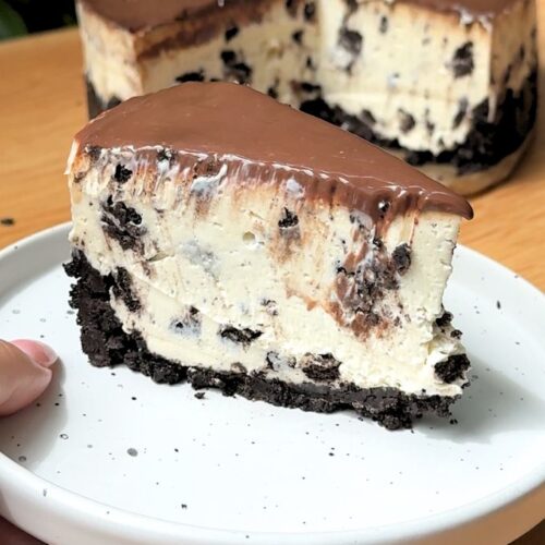 A single slice of no bake Oreo cheesecake on a plate sitting right next to the main whole cheesecake.
