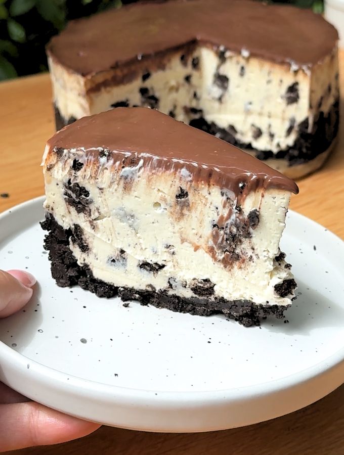 A single slice of no bake Oreo cheesecake on a plate sitting right next to the main whole cheesecake.