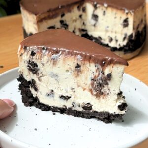 A single slice of no bake Oreo cheesecake on a plate sitting right next to the main whole cheesecake.