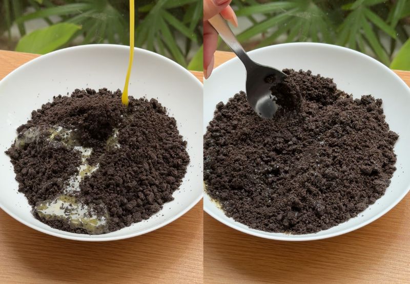 A hand using a rubber spatula to mix dark cookie crumbs and melted butter in a bowl.