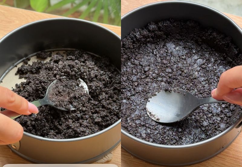 A hand using the flat bottom of a measuring cup to press chocolate cookie crumbs firmly into a silver springform pan.