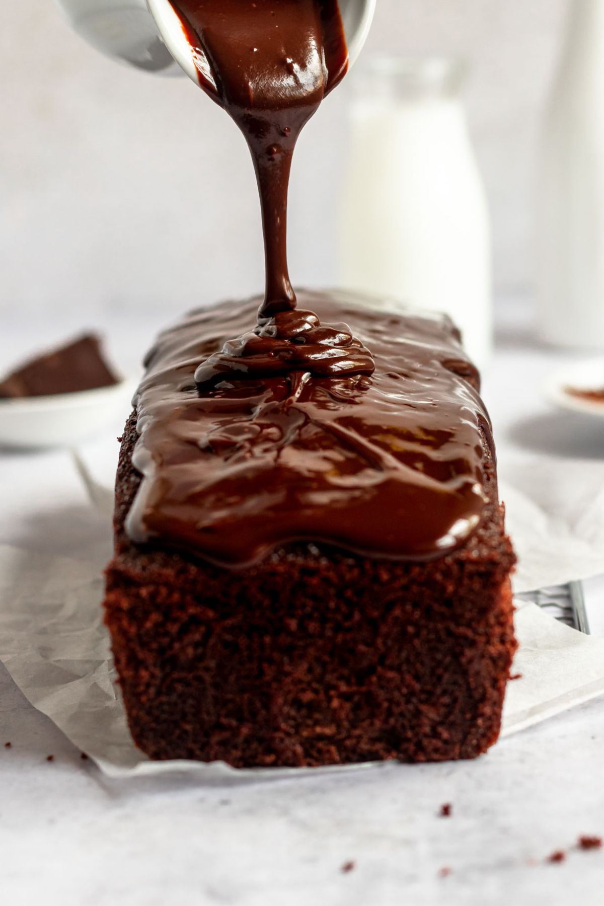 Smooth chocolate ganache being poured from a white pitcher onto a freshly baked chocolate pound loaf cake.