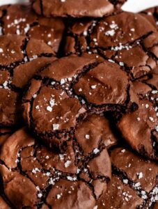 Overhead close-up of a pile of fudgy brownie cookies with crinkled tops and flaky sea salt, featuring a bite taken out of the center cookie.