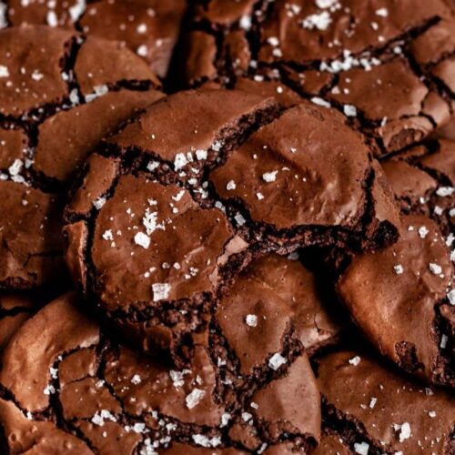 Overhead close-up of a pile of fudgy brownie cookies with crinkled tops and flaky sea salt, featuring a bite taken out of the center cookie.