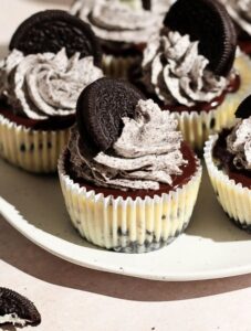Mini Oreo cheesecakes on a white plate topped with chocolate ganache and Oreo whipped cream.