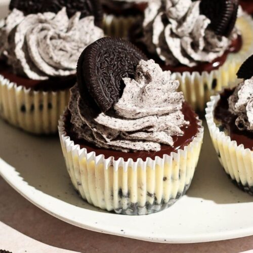 Mini Oreo cheesecakes on a white plate topped with chocolate ganache and Oreo whipped cream.