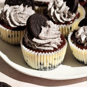 Mini Oreo cheesecakes on a white plate topped with chocolate ganache and Oreo whipped cream.