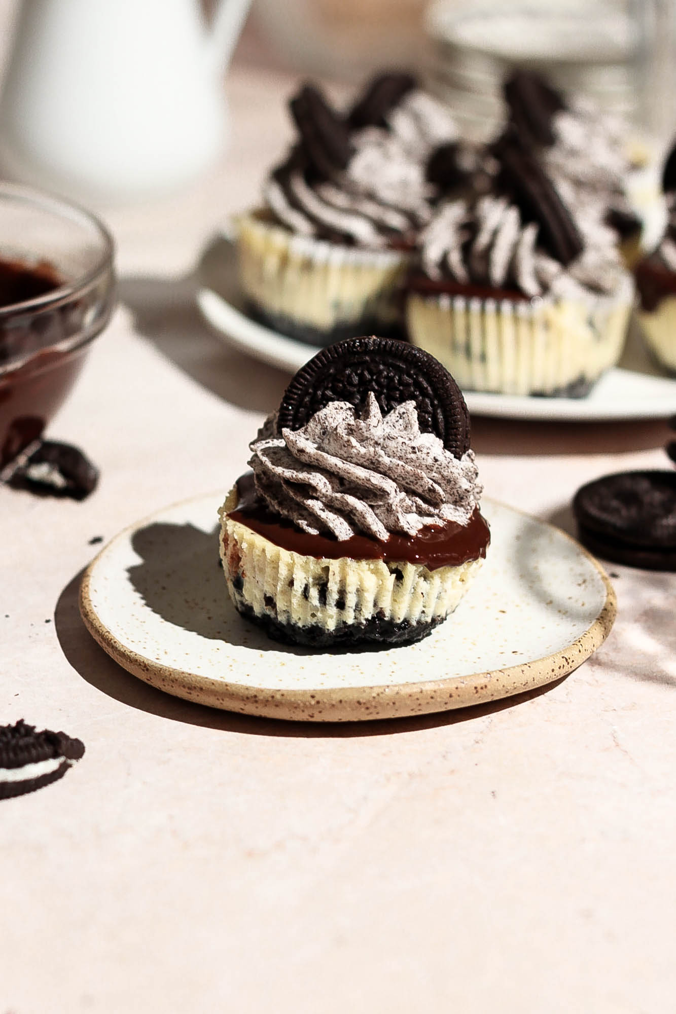 A single mini Oreo cheesecake served on a small plate with a bowl of chocolate ganache in the background