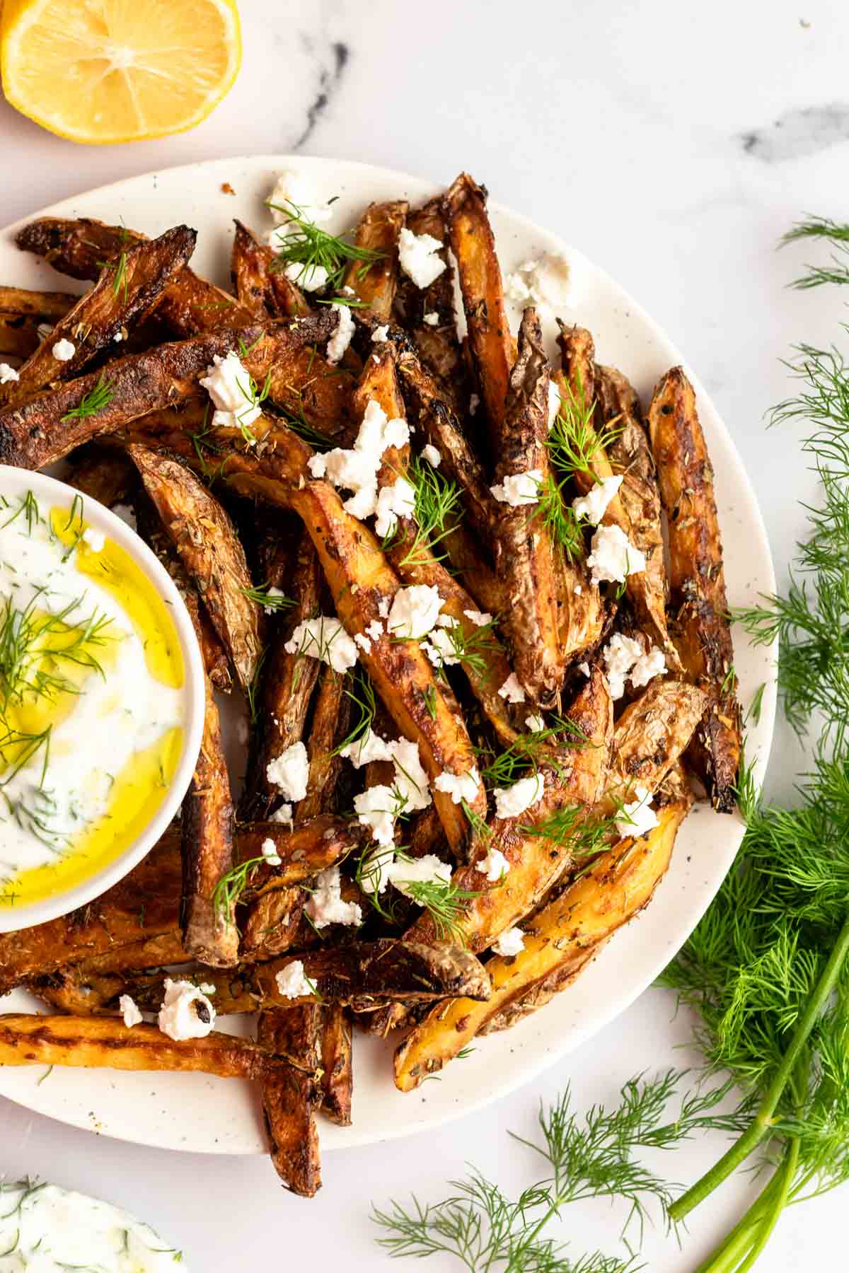 Large plate filled with baked greek fries next to a bowl with yogurt dill sauce.