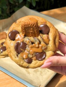 A hand holding up a whole, golden-brown Biscoff butter cookie topped with crushed Lotus biscuit crumbs.