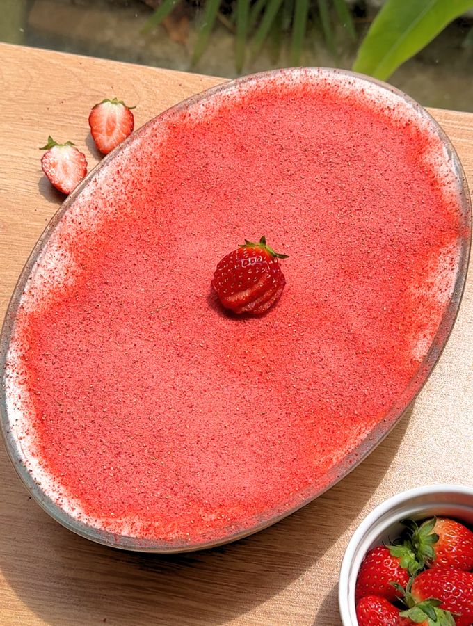 A close-up shot of a beautiful oval baking dish filled with a full strawberry tiramisu, topped with red strawberry powder and fresh sliced strawberries.