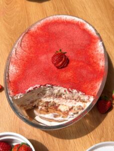 A top-down shot of a strawberry tiramisu in an oval baking dish with one slice cut out, showing the layered ladyfingers and pink cream inside the pan.