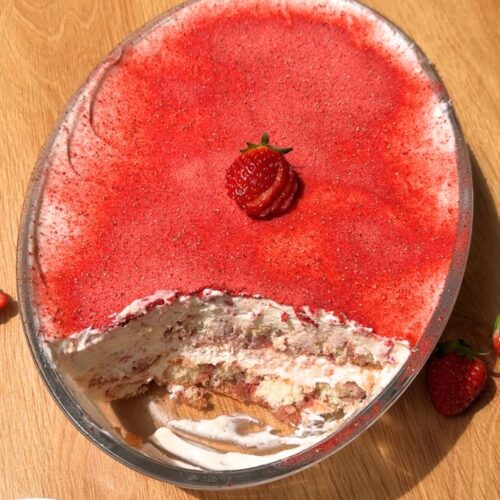 A top-down shot of a strawberry tiramisu in an oval baking dish with one slice cut out, showing the layered ladyfingers and pink cream inside the pan.