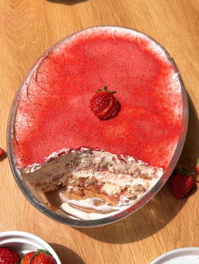A top-down shot of a strawberry tiramisu in an oval baking dish with one slice cut out, showing the layered ladyfingers and pink cream inside the pan.