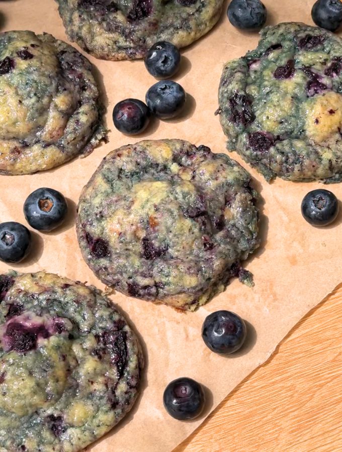 Blueberry cheesecake cookies on a brown parchment paper next to fresh blueberries.