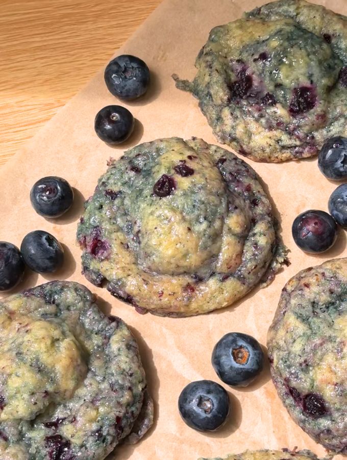 Blueberry cheesecake cookies on a brown parchment paper next to fresh blueberries.