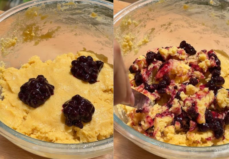 A hand using a silver butter knife to gently drag through the cookie dough and blueberry jam in a zig-zag pattern, creating distinct purple ribbons.