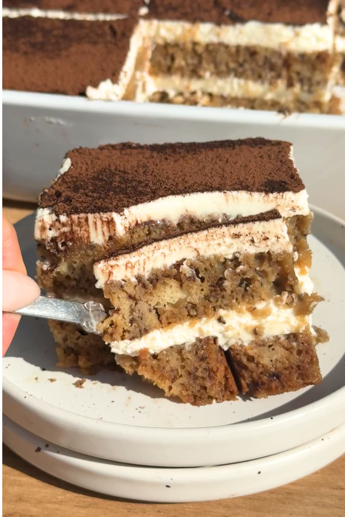 A fork cutting a bite from a slice of layered banana bread tiramisu dusted with cocoa powder.
