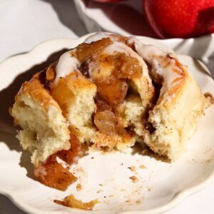 Apple cinnamon roll with cream cheese frosting and chunky apple filling on a white plate, with cinnamon sticks nearby.