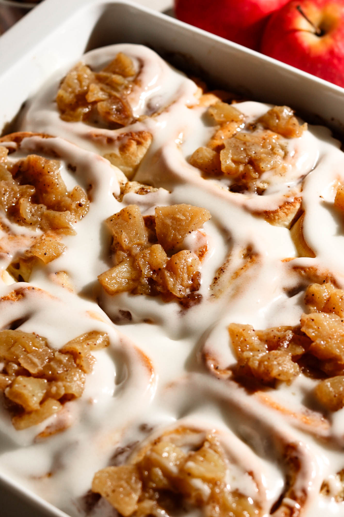 Close-up of baked apple cinnamon rolls in a white baking dish covered with white icing and chunky spiced apple topping, with fresh red apples in the background.