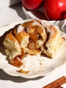 Apple cinnamon roll with cream cheese frosting and chunky apple filling on a white plate, with cinnamon sticks nearby.