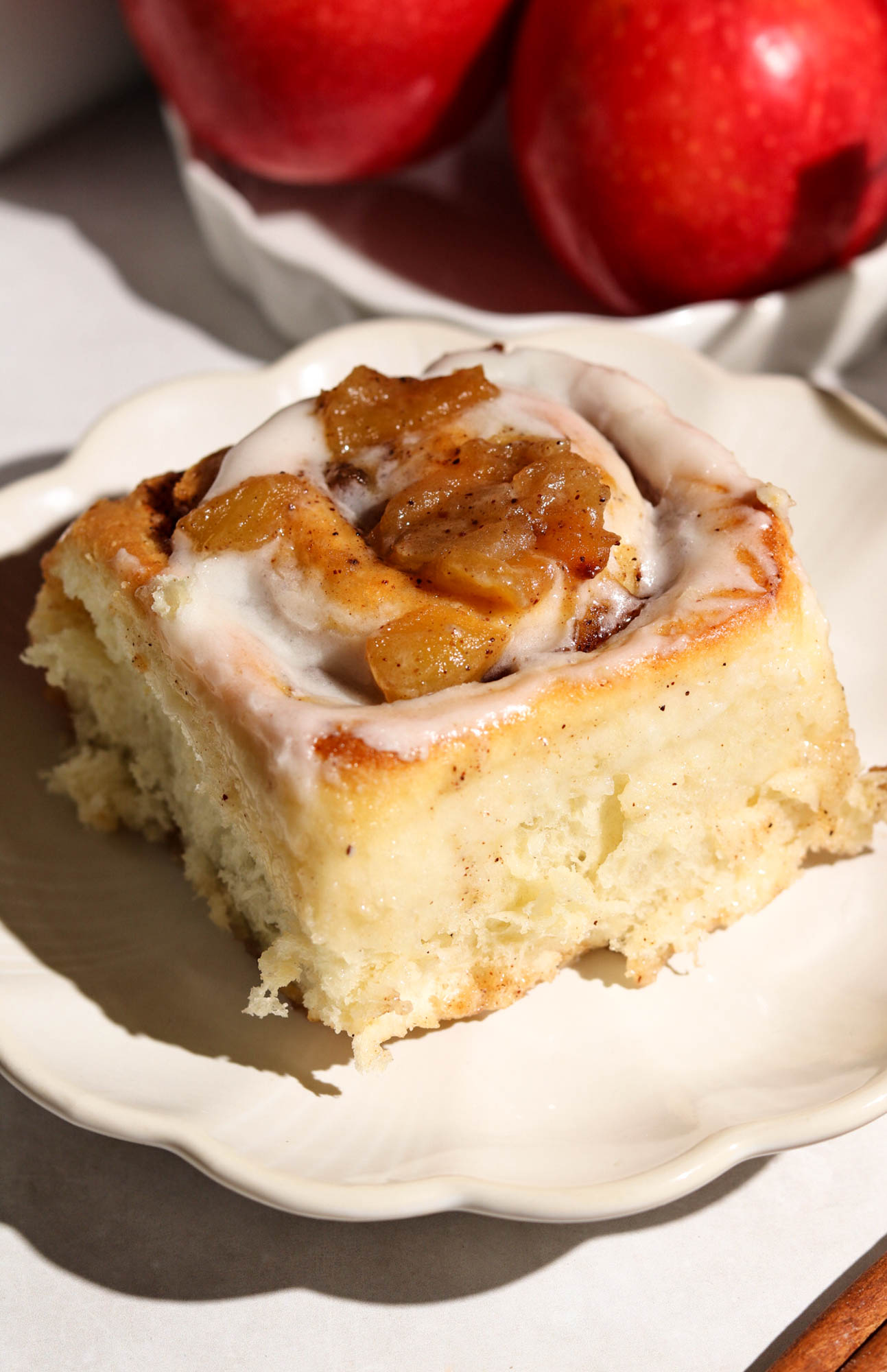 Close-up of a single apple cinnamon roll with white icing and apple chunks on a white plate, with red apples in the background.