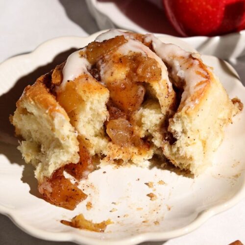 Apple cinnamon roll with cream cheese frosting and chunky apple filling on a white plate, with cinnamon sticks nearby.