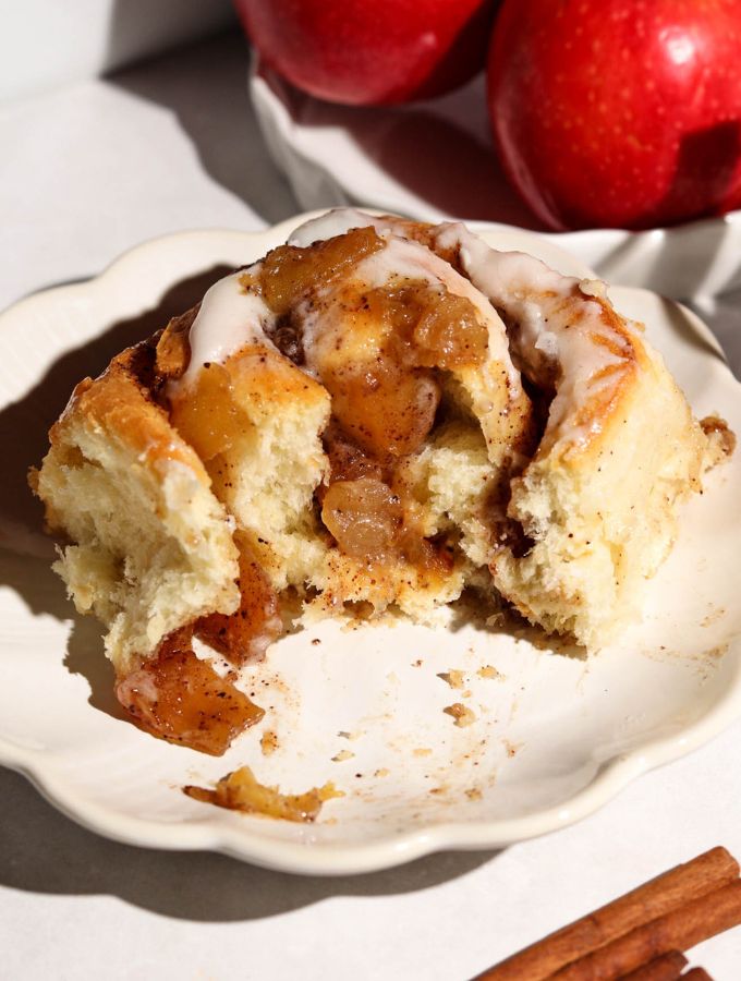 Apple cinnamon roll with cream cheese frosting and chunky apple filling on a white plate, with cinnamon sticks nearby.