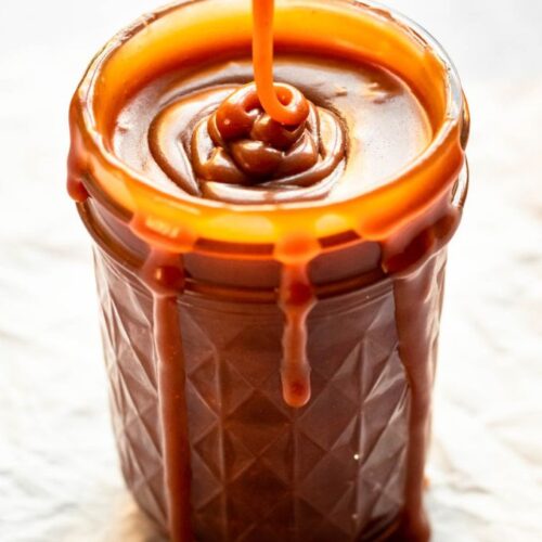 Rich salted caramel sauce pouring over the rim of a glass jar with thick, decadent drips running down the sides.