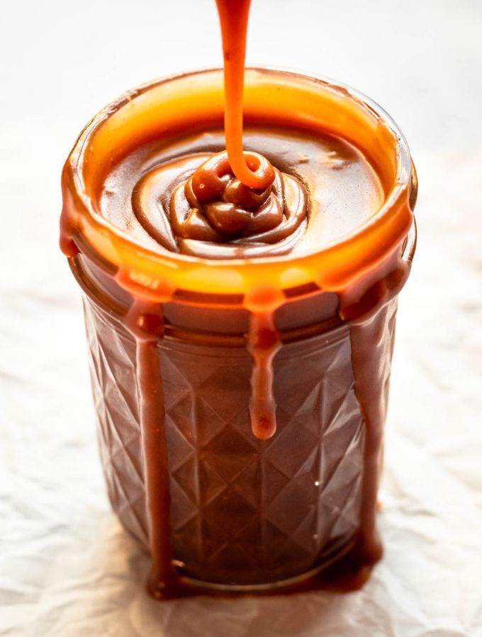 Rich salted caramel sauce pouring over the rim of a glass jar with thick, decadent drips running down the sides.