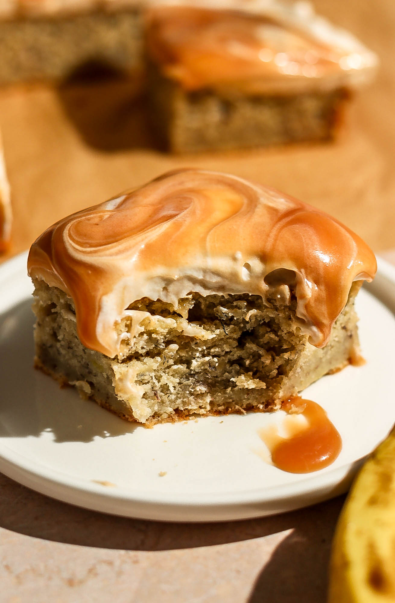 A close-up, angled shot of a single slice of caramel banana cake on a white plate with a bite taken, highlighting the dripping caramel syrup.