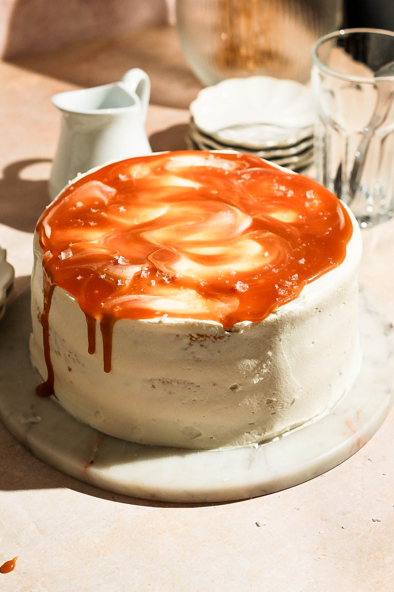 A frosted round cake topped with swirling salted caramel, flaky sea salt, and caramel drips on a marble stand.